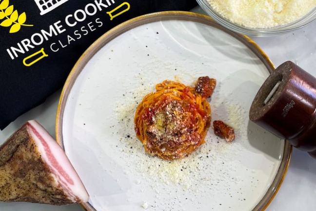Plate of spaghetti with cheese, surrounded by cheese bowl, meat, and pepper mill. Cooking class apron nearby.