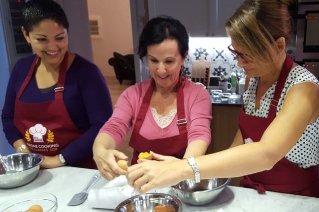 Three women cracking eggs into bowls, wearing aprons and smiling in a kitchen.